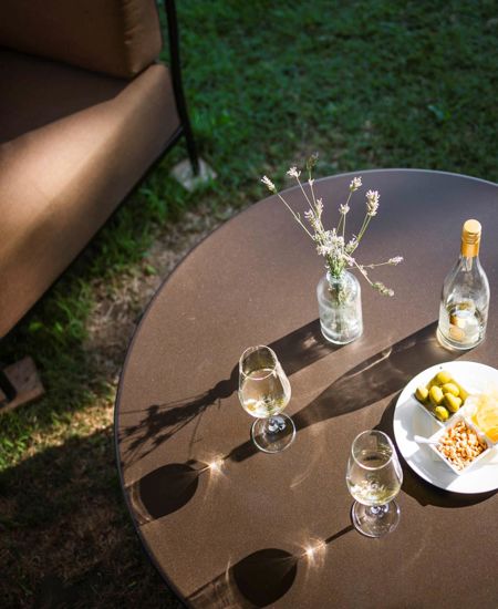 Ein Tisch im Garten von oben. Auf dem Tisch befinden sich zwei Gläser mit Weißwein, die Flasche, eine Platte mit Chips, Erdnüssen und Oliven, sowie die Vase mit Blüten.